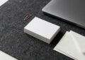 Pile de cartes de visite vierges et enveloppes blanches sur un sous-main en feutre gris, à côté d’un ordinateur portable fermé
