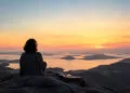Accueil 51 Une personne assise sur une falaise avec un casque et une guitare, contemplant un coucher de soleil sur la mer.