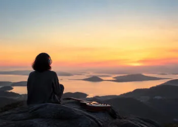 Suno AI avis : ce que vaut le générateur de chansons en 2026 1 Une personne assise sur une falaise avec un casque et une guitare, contemplant un coucher de soleil sur la mer.