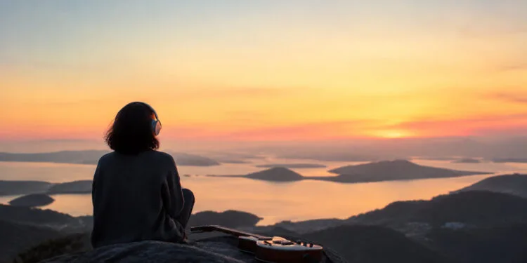 Accueil 43 Une personne assise sur une falaise avec un casque et une guitare, contemplant un coucher de soleil sur la mer.