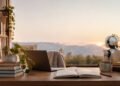 Un petit robot blanc assis à un bureau avec un ordinateur portable, des livres et une vue sur les montagnes au coucher.