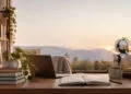 Accueil 54 Un petit robot blanc assis à un bureau avec un ordinateur portable, des livres et une vue sur les montagnes au coucher.