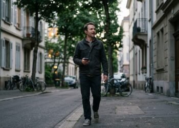 Homme qui marche dans une rue arborée en parlant à voix haute, écouteurs sans fil et téléphone en main